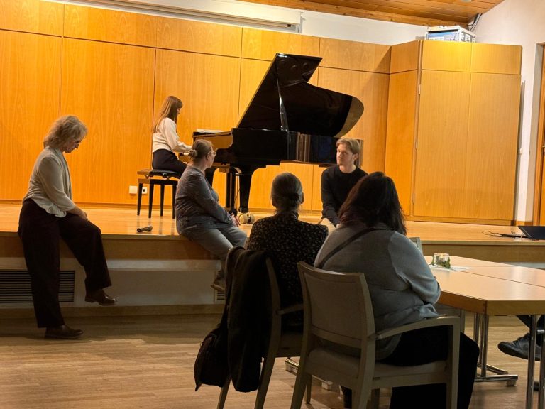 Eine Person spielt Klavier, während andere aufmerksam zuhören.