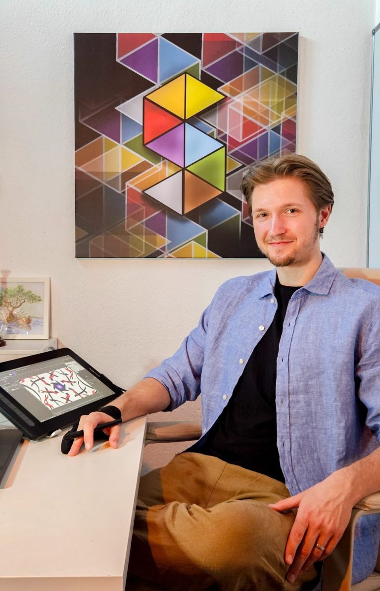 Der Künstler sitzt an einem Schreibtisch mit Tablet und vor einem bunten geometrischen Wandbild.